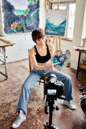 Téléchargez les photos : Une femme s'assoit sur une chaise, tenant une peinture avec une expression réfléchie. - en image libre de droit