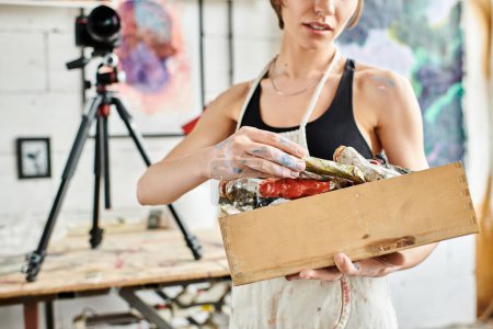 Mujer con confianza sostiene una caja de pintura delante de una cámara.