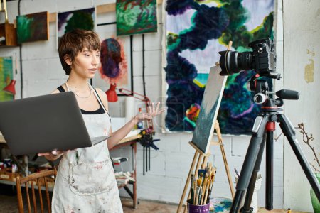 Woman in art studio holding a laptop, surrounded by creativity.