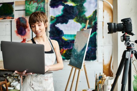 A woman holds a laptop in front of a painting.