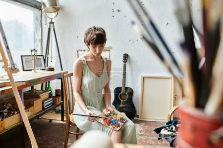 A woman sits in a room with a guitar, holding brush and palette.