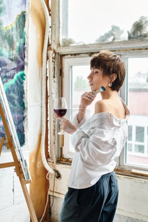 A woman stands by a window, holding a glass of wine.