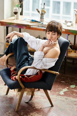 Young woman with dapper style sits in a chair, smoking a cigarette in art studio.