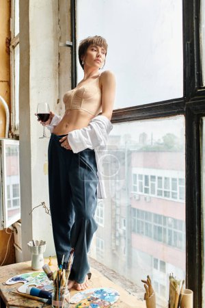 Eine stilvolle Frau hält ein Glas Wein, während sie vor einem Fenster steht.