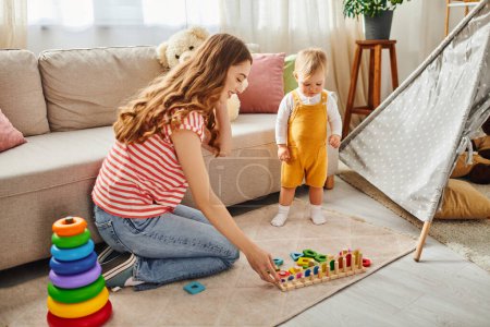 Téléchargez les photos : Une jeune mère interagit joyeusement avec sa fille tout-petit tout en jouant sur le sol à la maison. - en image libre de droit