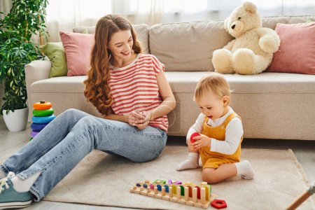Eine junge Mutter sitzt auf dem Boden und lässt sich durch spielerische Interaktionen freudig auf ihre kleine Tochter ein.
