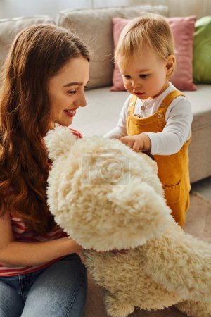 Téléchargez les photos : Une jeune mère tient avec amour un animal en peluche à côté de sa fille tout-petit à la maison, créant un moment chaleureux d'affection. - en image libre de droit