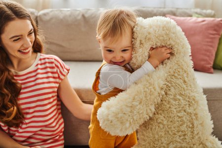 Eine junge Mutter hält ihr Baby zärtlich in der Hand, während sie einen Teddybär in einer herzerwärmenden Liebeserklärung zu Hause umarmt.