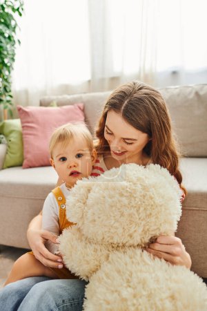 A young mother embraces her baby daughter while holding a teddy bear, enjoying a precious moment of bonding and love at home.