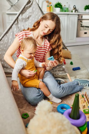 Téléchargez les photos : Une jeune mère s'assoit sur le sol, jouant joyeusement avec sa petite fille, créant un moment chaleureux d'amour et de connexion. - en image libre de droit