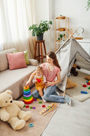 Une petite fille rit en jouant avec son ours en peluche dans une tente de tipi, créant des souvenirs magiques avec sa jeune mère.