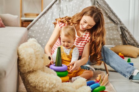 Téléchargez les photos : A young mother and her toddler daughter happily playing with a teddy bear, creating heartwarming memories at home. - en image libre de droit