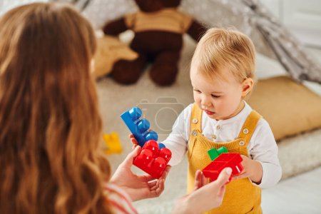 A young mother lovingly holds her baby daughter as they joyfully play with colorful toys together at home.