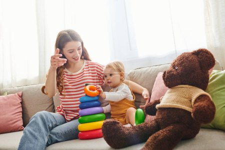 A young mother sits on a couch with her baby daughter and a teddy bear, sharing a tender moment of love and togetherness.
