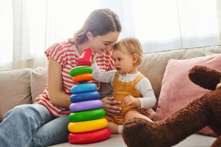 Eine junge Mutter und ihre kleine Tochter lachen und spielen zusammen auf einer Couch in einer gemütlichen häuslichen Umgebung.