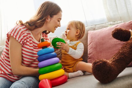 A young mother joyfully playing with her toddler daughter on a cozy couch at home.