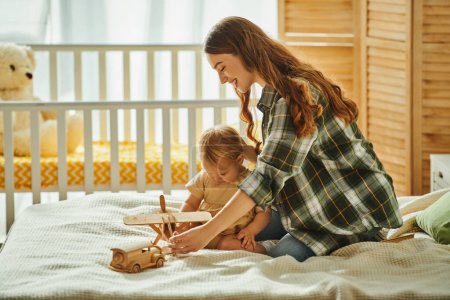 Foto de Una joven madre juega alegremente con su hija pequeña en una cama acogedora, compartiendo sonrisas y risas en una escena conmovedora. - Imagen libre de derechos