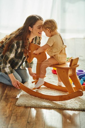 A young mother joyfully playing with her toddler daughter on a rocking horse at home.