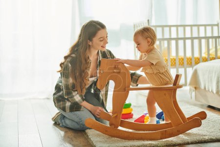 A young mother joyfully plays with her toddler daughter on a wooden rocking horse in their cozy home.