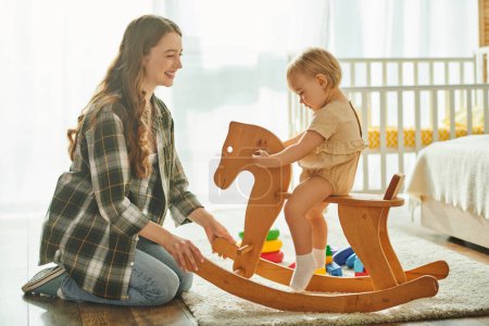 Une jeune mère joue joyeusement avec sa fille tout-petit sur un cheval à bascule à la maison, collant et créant des souvenirs spéciaux ensemble.