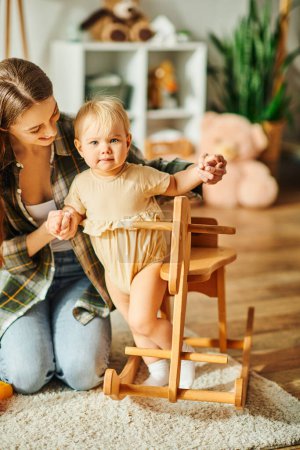 A young mother kneels next to her toddler daughter sitting in a high chair, sharing a precious moment together at home.