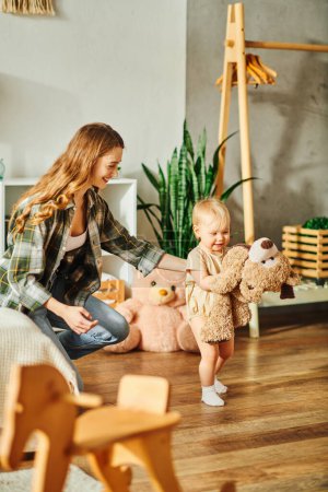A young mother laughing as she plays with her baby girl and a teddy bear on the floor at home.