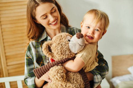 Eine junge Mutter hält ihre kleine Tochter auf dem Arm, während das Kleinkind einen Teddybär streichelt und damit Liebe und Verbundenheit zeigt.