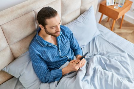 Foto de A man peacefully sits on a bed, focused on his cell phone screen. - Imagen libre de derechos