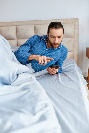 A man lies in bed, absorbed by his cell phone screen.