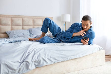 Schöner Mann, der auf dem Bett liegt und Yoga praktiziert, während er sein Handy hält.