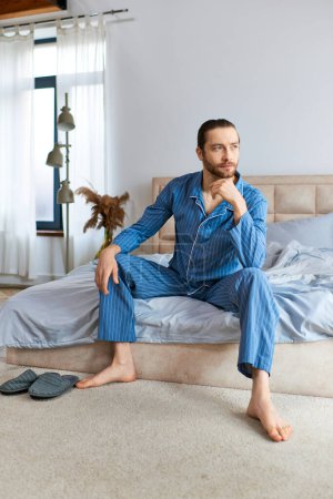A handsome man in pajamas sits calmly on a bed.