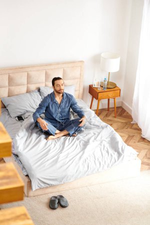 Téléchargez les photos : Beau homme pratique le yoga tout en étant assis sur le lit dans une chambre confortable. - en image libre de droit
