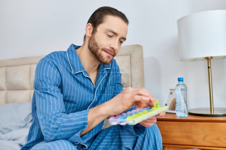 Handsome man sits on bed, holding holding pill case in a calm and serene manner.