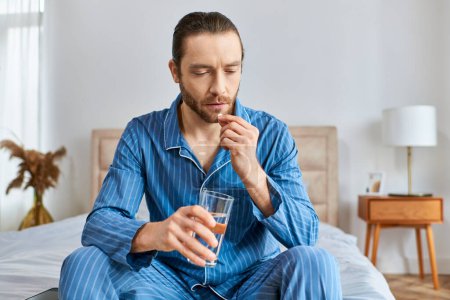 Foto de Un hombre sentado en una cama, bebiendo tranquilamente un vaso de agua con una pastilla. - Imagen libre de derechos