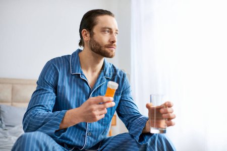 Photo for Handsome man sitting on bed, holding a bottle of pills and glass. - Royalty Free Image