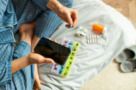A man holding a smart phone and pill in bed in the morning.