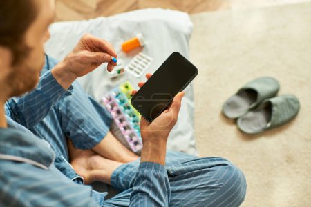 A man seated on the floor engages with his smartphone and pills.