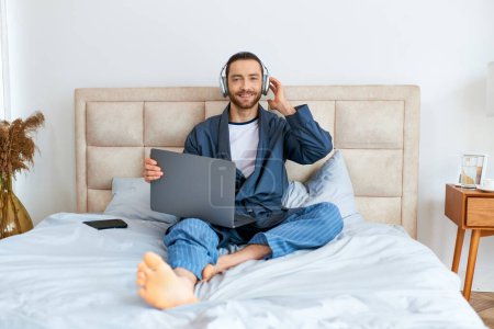 Photo for A man sitting on a bed, using a laptop and wearing headphones. - Royalty Free Image