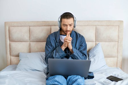 Foto de Un hombre sentado en una cama, usando auriculares, usando una computadora portátil. - Imagen libre de derechos