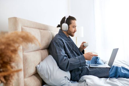 Téléchargez les photos : Un homme au lit avec un ordinateur portable et un casque immergé dans sa routine matinale. - en image libre de droit