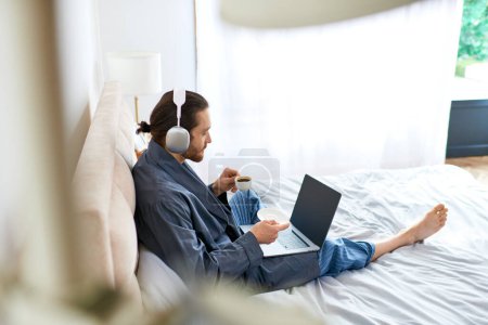 Téléchargez les photos : Homme assis sur le lit, en utilisant un ordinateur portable pour la relaxation matinale. - en image libre de droit