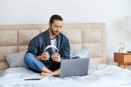 Foto de Un hombre mira la pantalla de un portátil mientras está sentado en una cama en un entorno tranquilo por la mañana. - Imagen libre de derechos