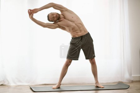 Photo for Handsome man standing on yoga mat, practicing yoga in front of window. - Royalty Free Image