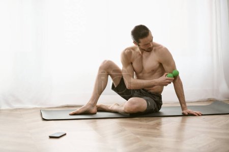 Photo for Shirtless man sits on yoga mat, holding a green massage ball. - Royalty Free Image
