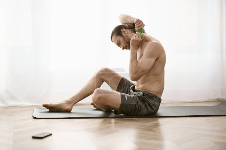 A handsome man peacefully sits shirtless on a yoga mat.