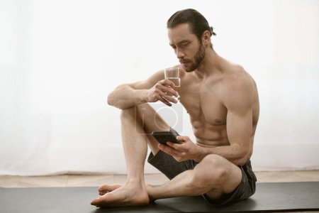 Un hombre sin camisa sentado en una esterilla de yoga, mirando el teléfono celular.
