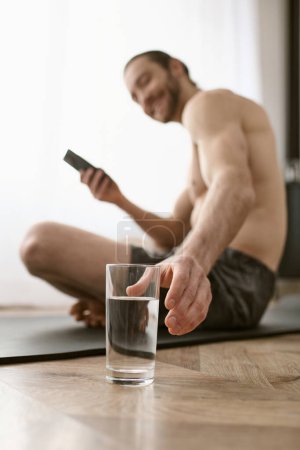 Un homme assis sur le sol, tenant un verre d'eau et un téléphone portable.