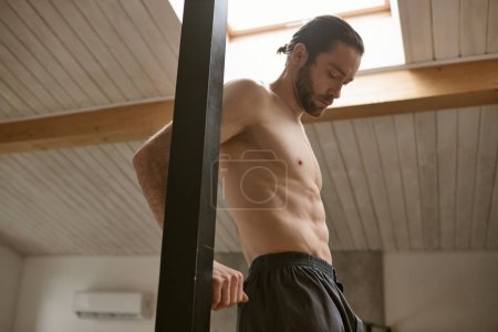 A shirtless man stands in a room with a pole, engaging in his energizing morning routine.