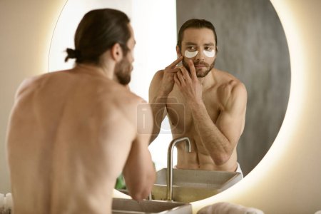 Foto de Un hombre guapo aplicando parches delante de un espejo durante su rutina de cuidado de la piel matutina. - Imagen libre de derechos