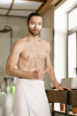 Hombre guapo con toalla alrededor de la cintura lleva a cabo rutina de cuidado de la piel por la mañana.